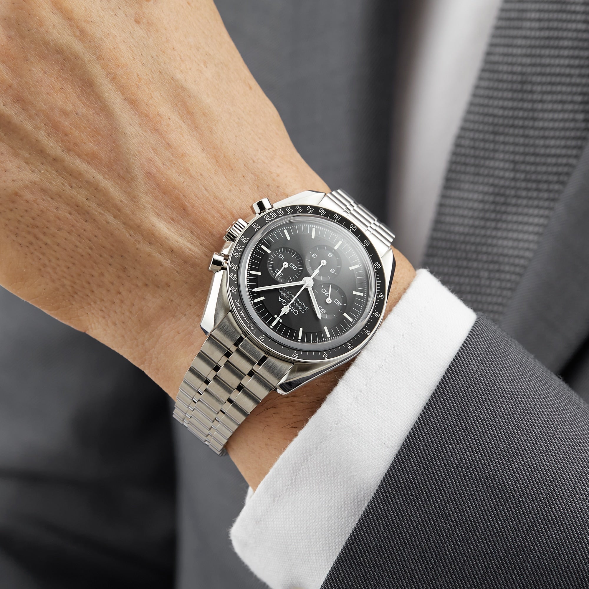 A person in a gray suit with a white shirt is wearing an OMEGA Speedmaster Moonwatch Professional, notable for its black dial, three subdials, and Master Chronometer certification.