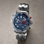 An OMEGA Seamaster Diver 300M Americas Cup Edition wristwatch with a blue dial, white markers, red and blue accents, and a metal bracelet is displayed on a grey surface.