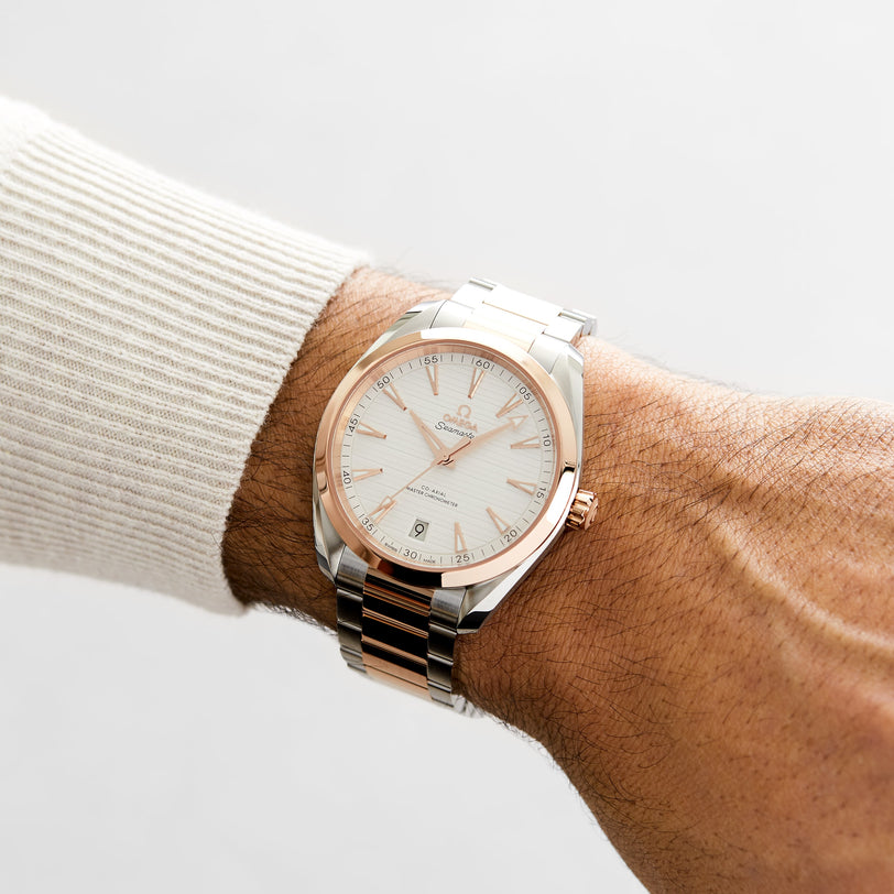 A person wears an OMEGA Seamaster Aqua Terra 150M wristwatch with a silver and Sedna™ gold band and a white dial on their left wrist, set against a plain background.