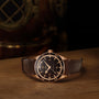 An OMEGA Seamaster 300 wristwatch with a gold case and brown leather strap rests on a wooden surface, with a brass diving helmet in the background.
