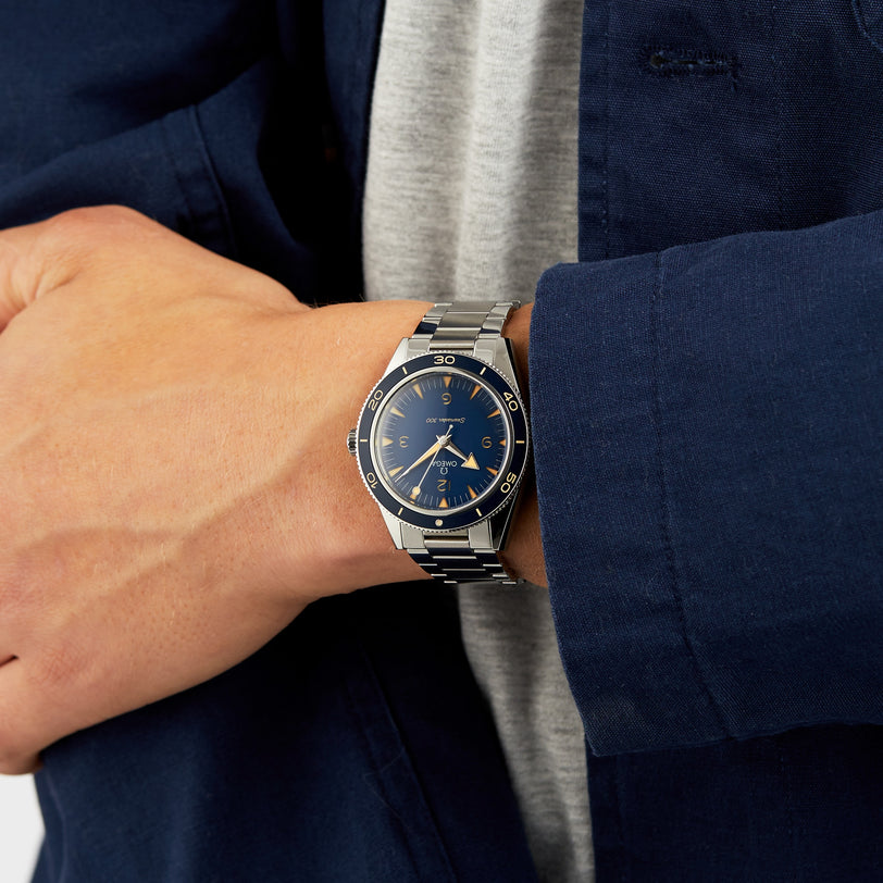 A person in a grey shirt and dark blue jacket wears an OMEGA Seamaster 300 dive watch with a blue face and metal band.