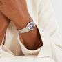 A person in a cream long-sleeve top rests their hands on a white surface, wearing an OMEGA Constellation watch with a silver case and round Patchouli blossom dial.
