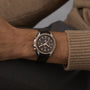 A close-up shows a person wearing an OMEGA Speedmaster Moonwatch Co-Axial Chronograph with a 42.5 mm stainless steel and 18K Sedna™ gold case, brown leather strap, and dark dial featuring a realistic moon image on their left wrist.