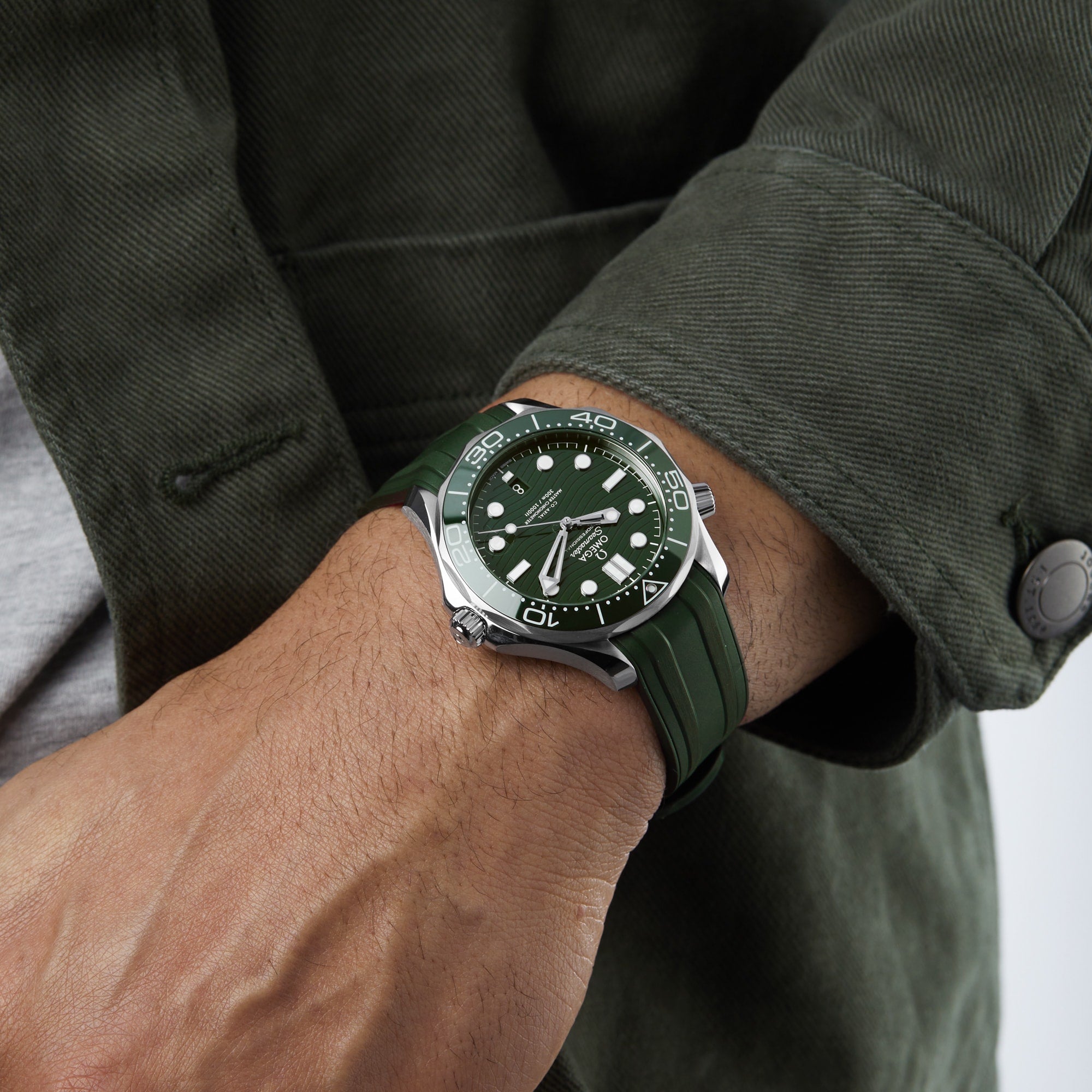 A person wears a green OMEGA Seamaster Diver 300M with a matching strap and jacket, their left hand on their right arm, highlighting the watchs precision and water resistance.