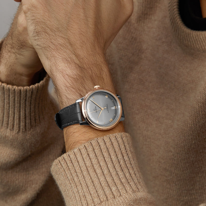 A person in a beige sweater showcases an OMEGA De Ville Prestige watch featuring a rose gold case, gray dial, and black leather strap on their left wrist.
