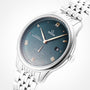 The OMEGA De Ville Prestige wristwatch features a silver case, blue dial with Roman numerals, date window at 3 o’clock, subdial at 6 o’clock, and a metal link bracelet, radiating refined elegance on a white background.