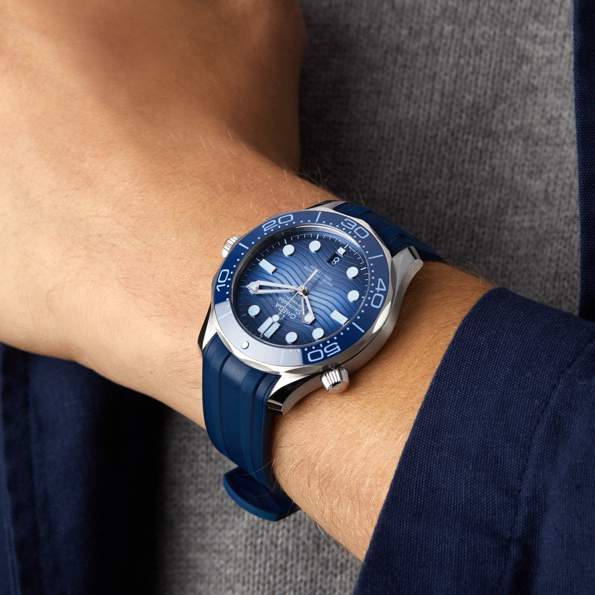 An individual sporting an OMEGA Seamaster Diver 300M Summer Blue with a silver bezel on their left wrist, paired with a grey shirt and navy blue jacket.