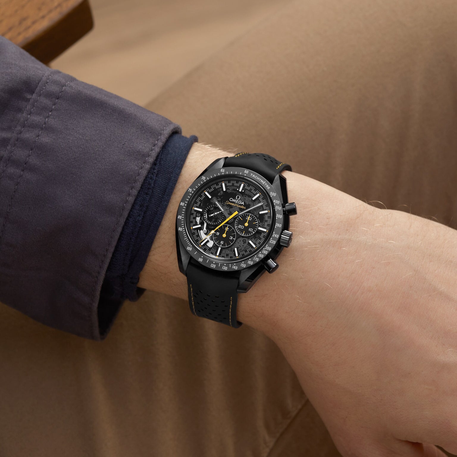 A person wearing an OMEGA Speedmaster Moonwatch Apollo 8 Dark Side Of The Moon with a dark strap, dressed in a gray jacket and tan pants, rests their arm on a surface.