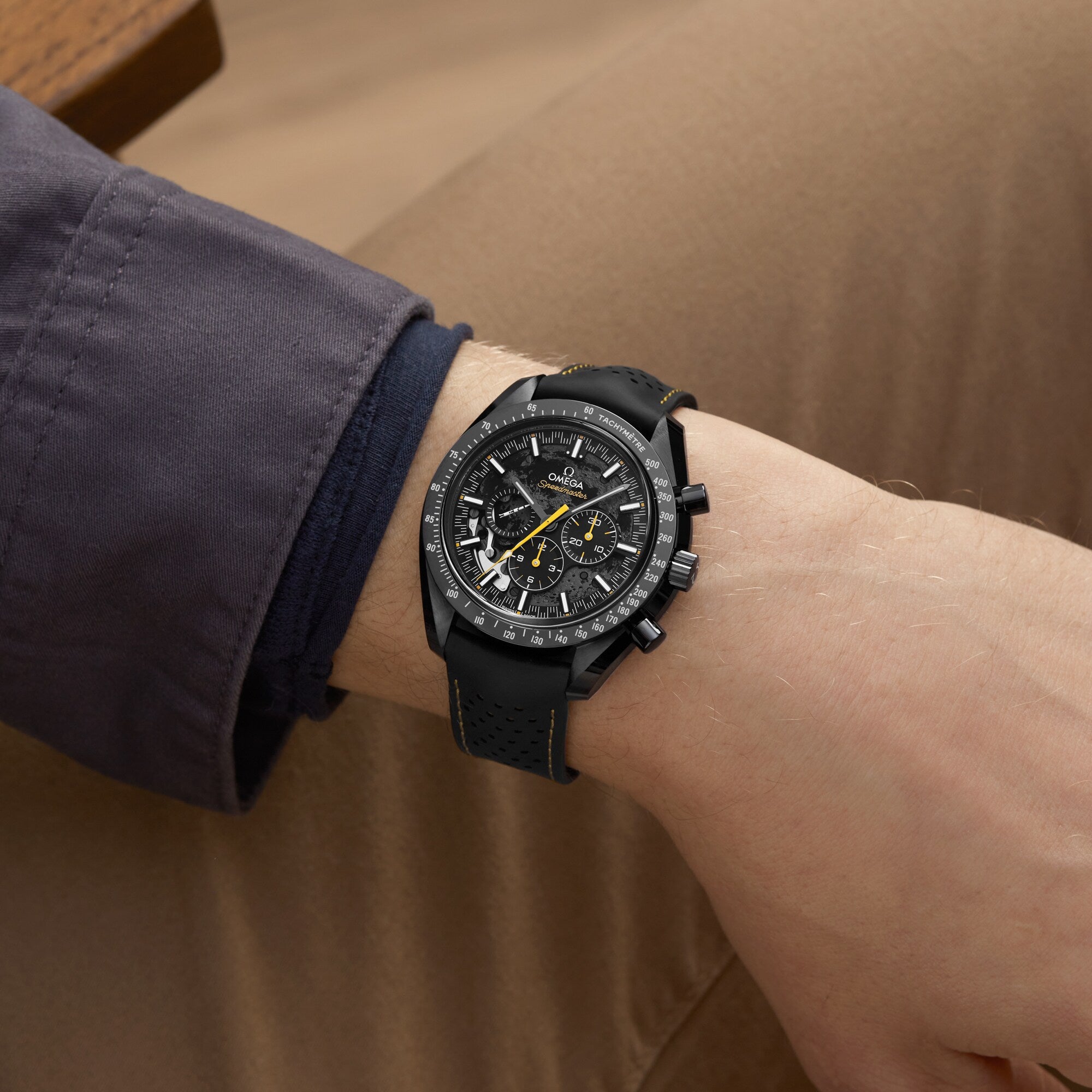 A person wearing an OMEGA Speedmaster Moonwatch Apollo 8 Dark Side Of The Moon with a dark strap, dressed in a gray jacket and tan pants, rests their arm on a surface.