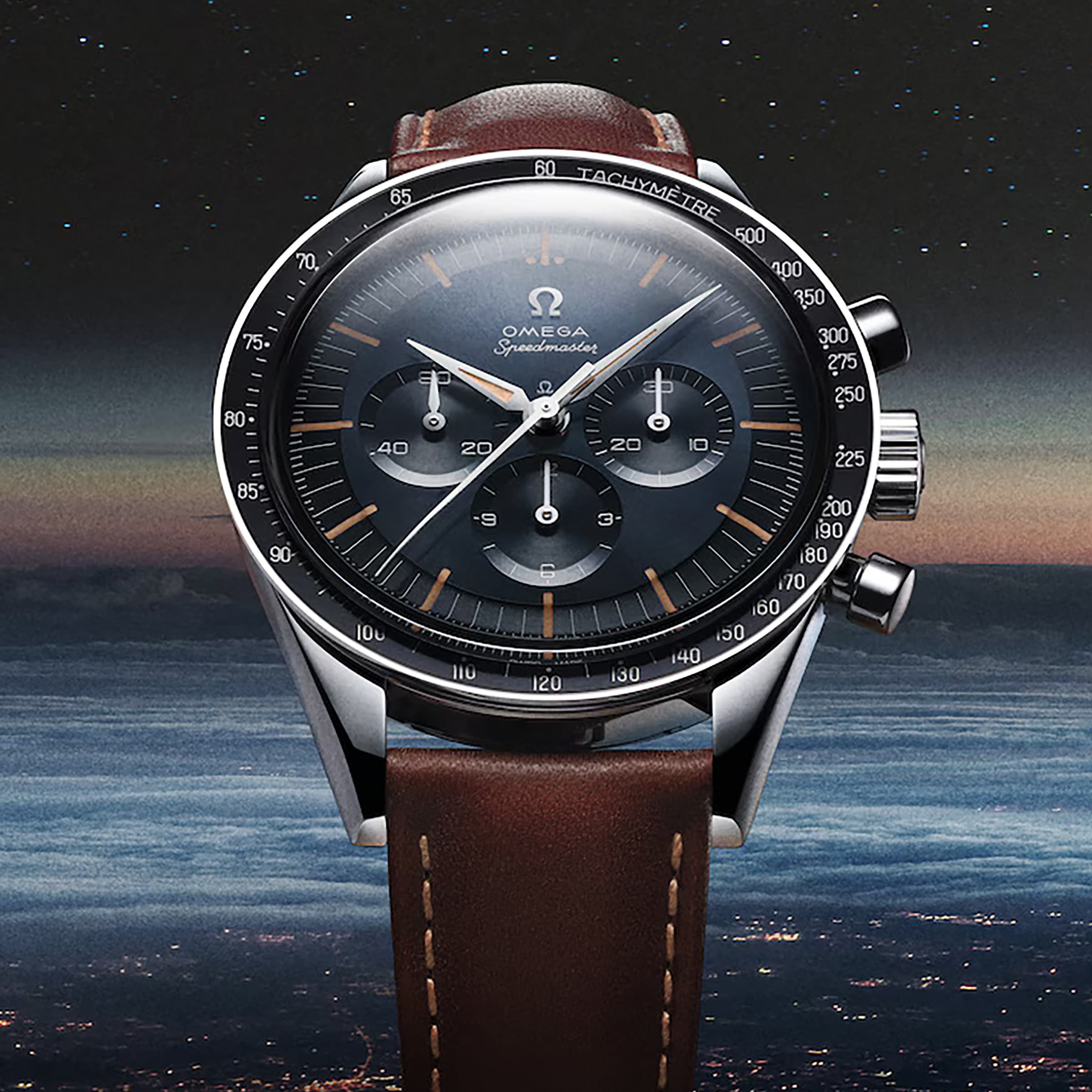A close-up of the OMEGA Speedmaster Anniversary Series chronograph watch with a brown leather strap and tachymeter scale, set against a night sky with a distant horizon.