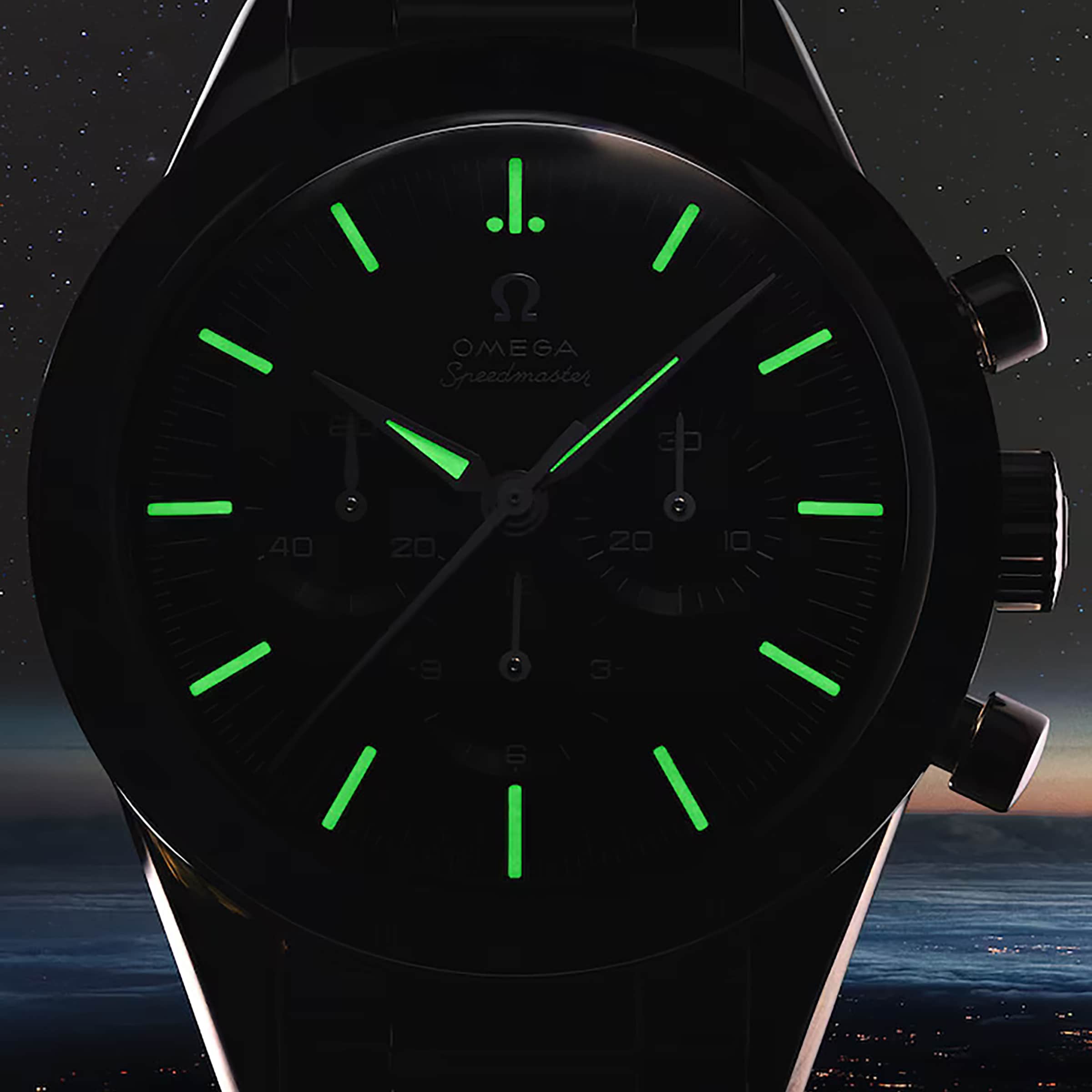 A close-up of an OMEGA Speedmaster Anniversary Series watch in darkness shows glowing green hour markers and hands on a faint dial, with the tachymeter scale subtly outlining the edge.