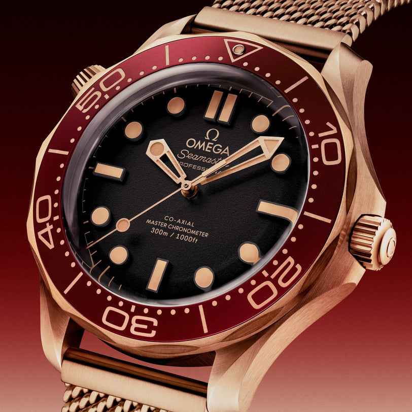 A close-up of the OMEGA Seamaster Diver 300M in Bronze Gold with a red bezel and mesh bracelet, showing the time at 10:10—a true Co-Axial Master Chronometer.