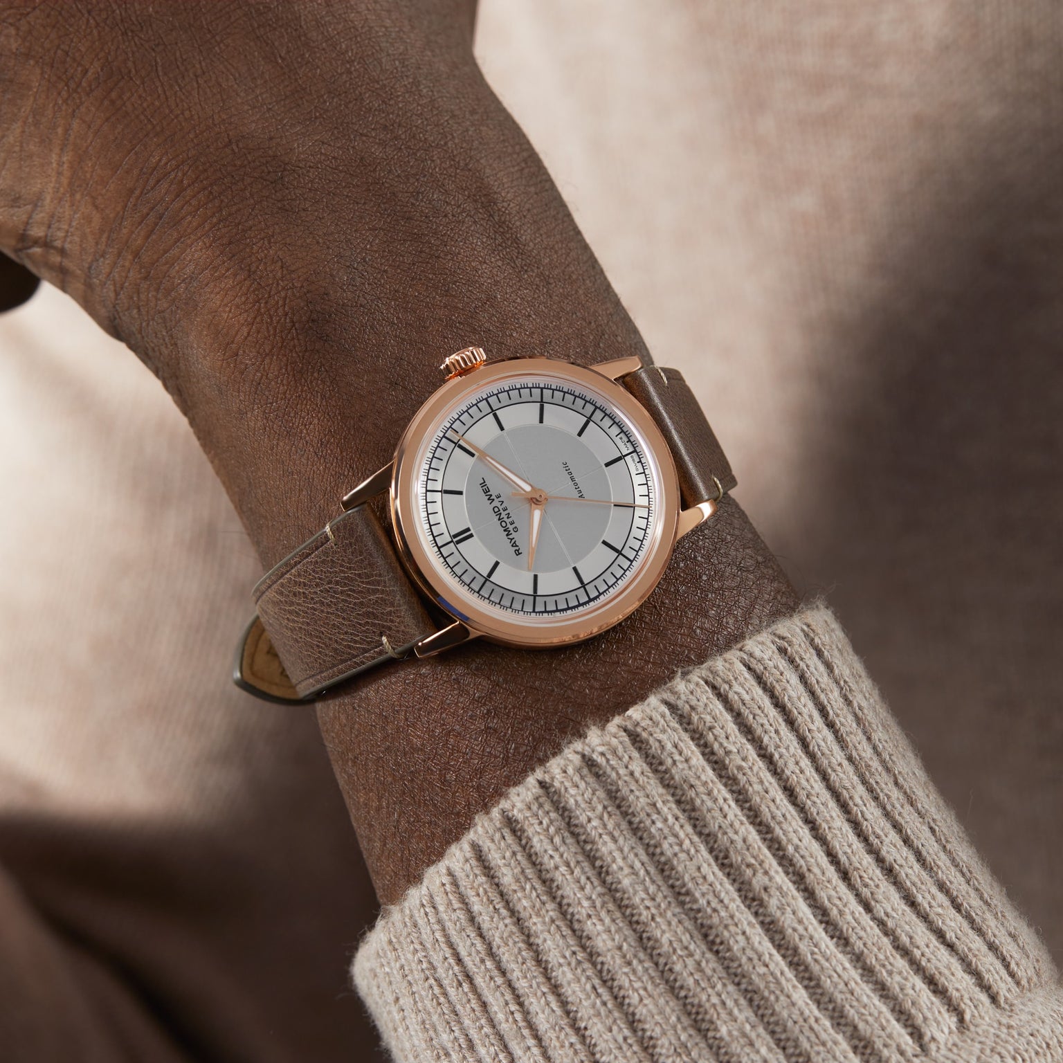 A close-up of a wrist in a beige sweater wearing the Raymond Weil Millesime Automatic Silver Sector Dial, a Swiss-made gold watch with a white face and beige leather strap.