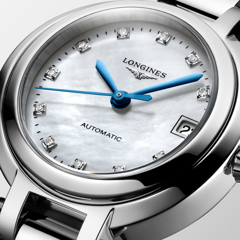 Close-up of the Longines PrimaLuna, a feminine automatic wristwatch with a white mother-of-pearl dial, blue hands, diamond-set markers, and a date window at 3 o’clock.