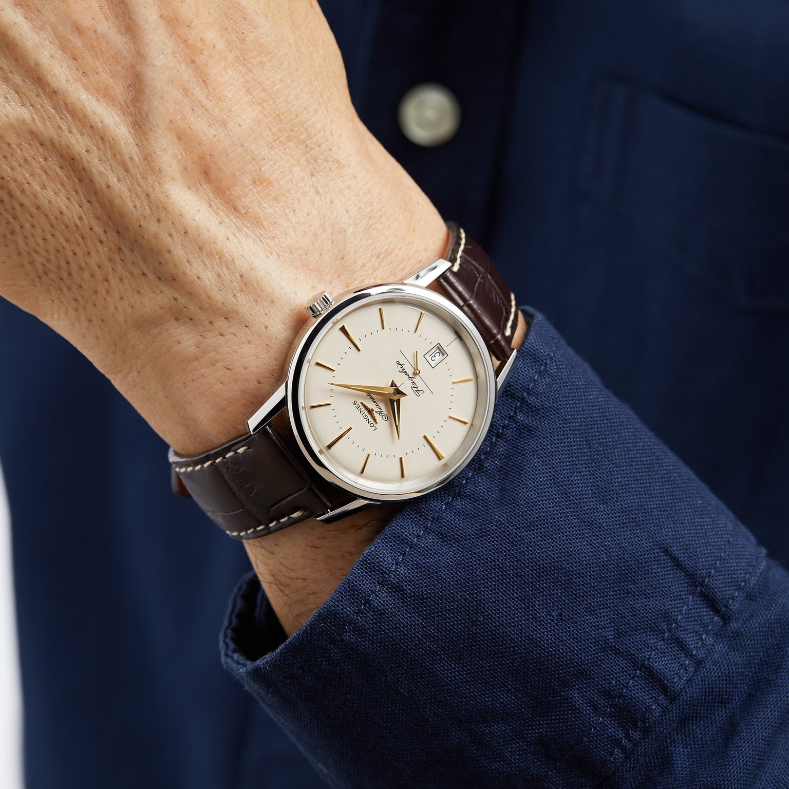 A person in a blue shirt wearing a Longines Flagship Heritage watch with a stainless steel case, brown leather strap, and cream-colored dial.