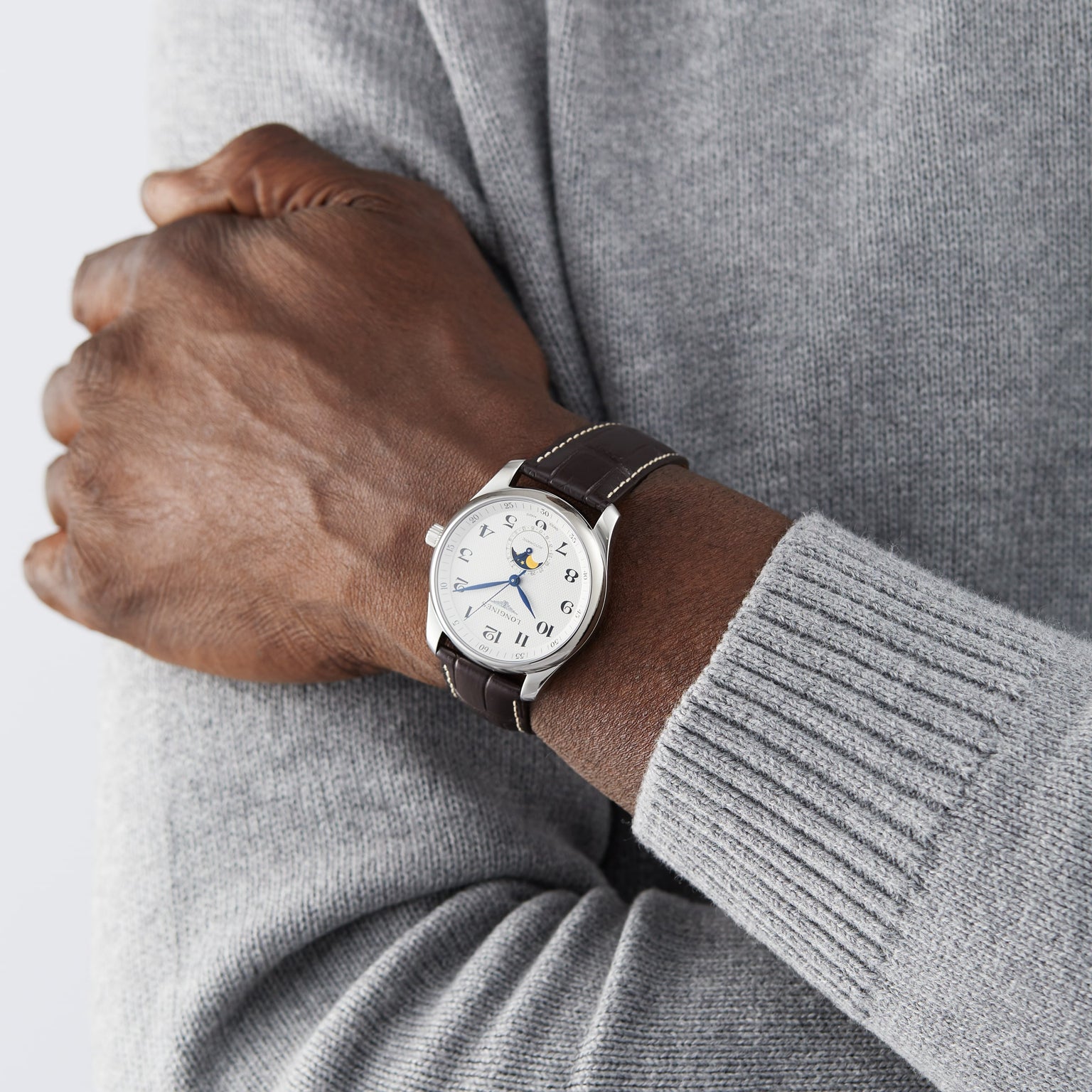 A person in a grey sweater crosses their arms, showcasing a Longines Master Collection Moonphase watch with a brown leather strap and white dial.