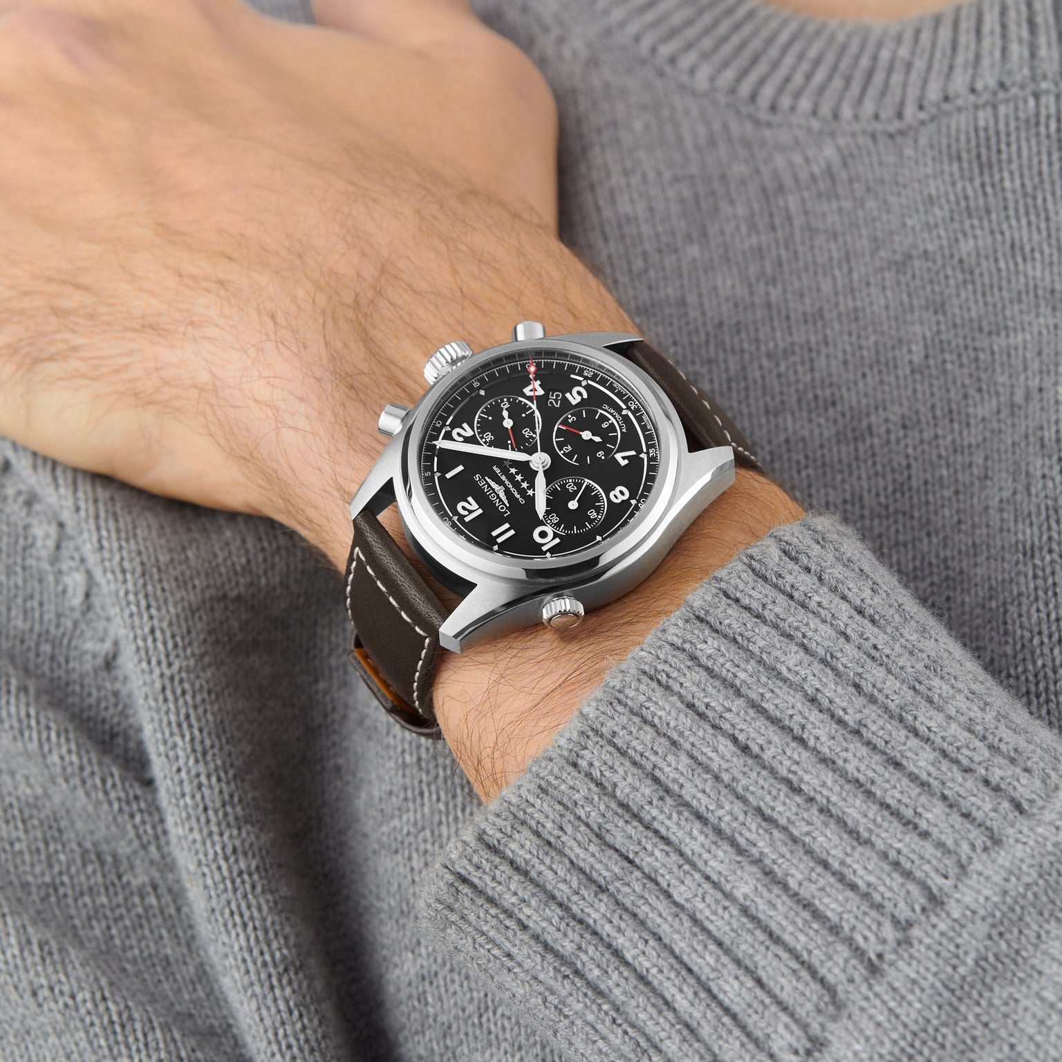 A person in a gray sweater showcases a Longines Spirit Chronograph with a black dial, chronograph subdials, dark leather strap, and a chronometer-certified movement on their left wrist.