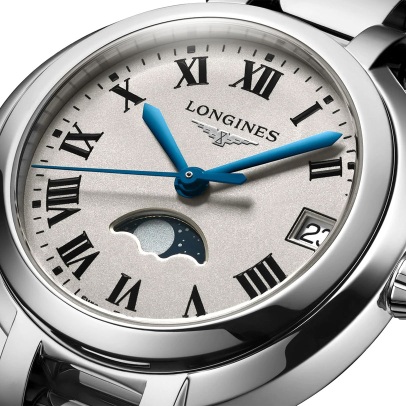 The Longines PrimaLuna Moonphase watch features Roman numerals, blue hands, a date display at 3 o’clock, and a moonphase at 6 o’clock on a silver dial set in a stainless steel case.
