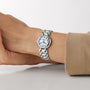 A person wears a Longines PrimaLuna Moonphase silver wristwatch with a white face, blue hands, Roman numerals, and a stainless steel case, resting their arm on a white surface.
