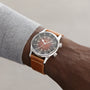 A person wears a Longines Legend Diver watch with a brown leather strap and black dial, showing 10:07. The watch pairs perfectly with their light gray long-sleeve shirt.