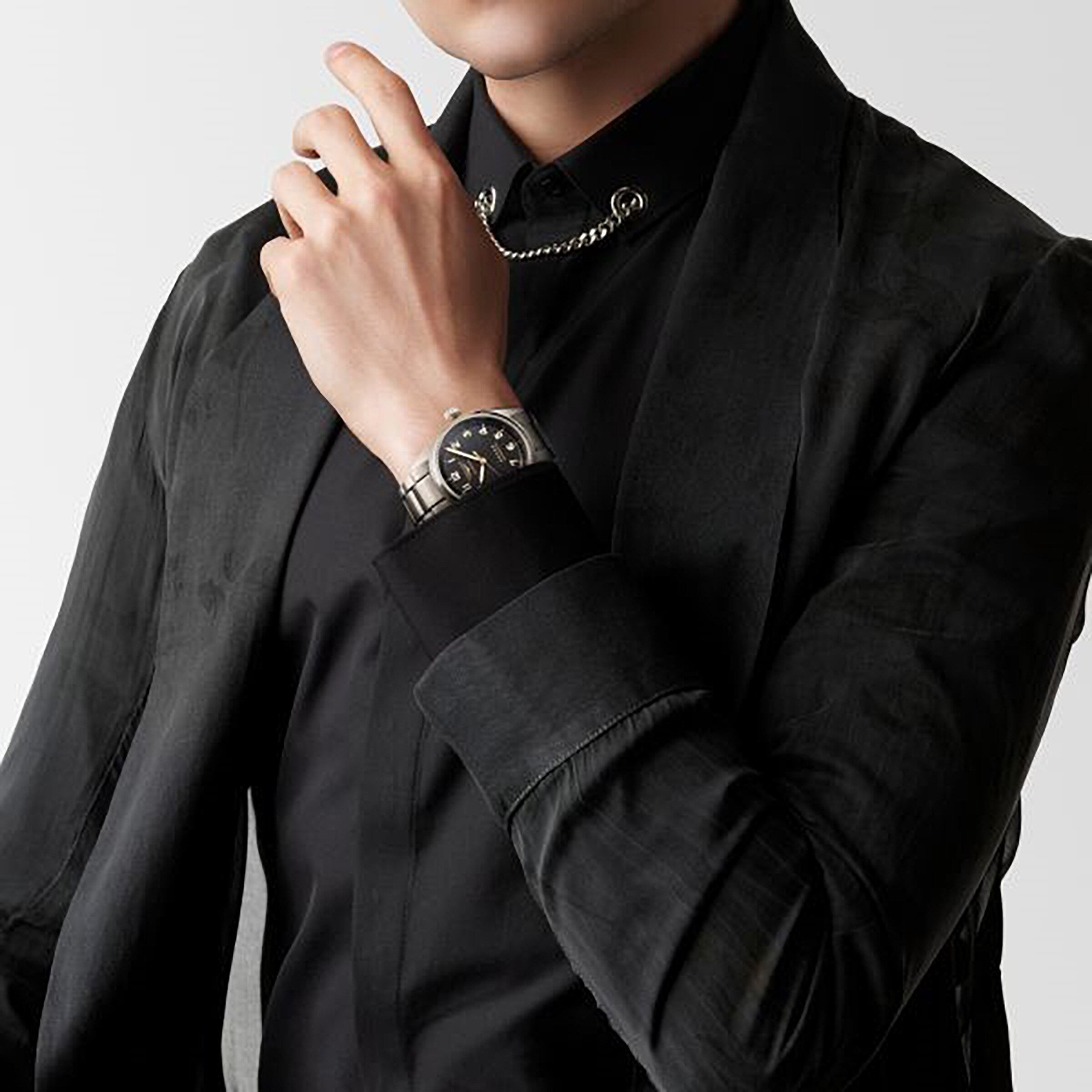 A person in a black shirt and jacket, wearing a silver chain necklace and a Longines Spirit watch, raises their hand near their face.