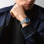 A man in a dark jacket rests his chin on his hand, wearing the Longines Spirit Zulu Time watch with a striking blue face.