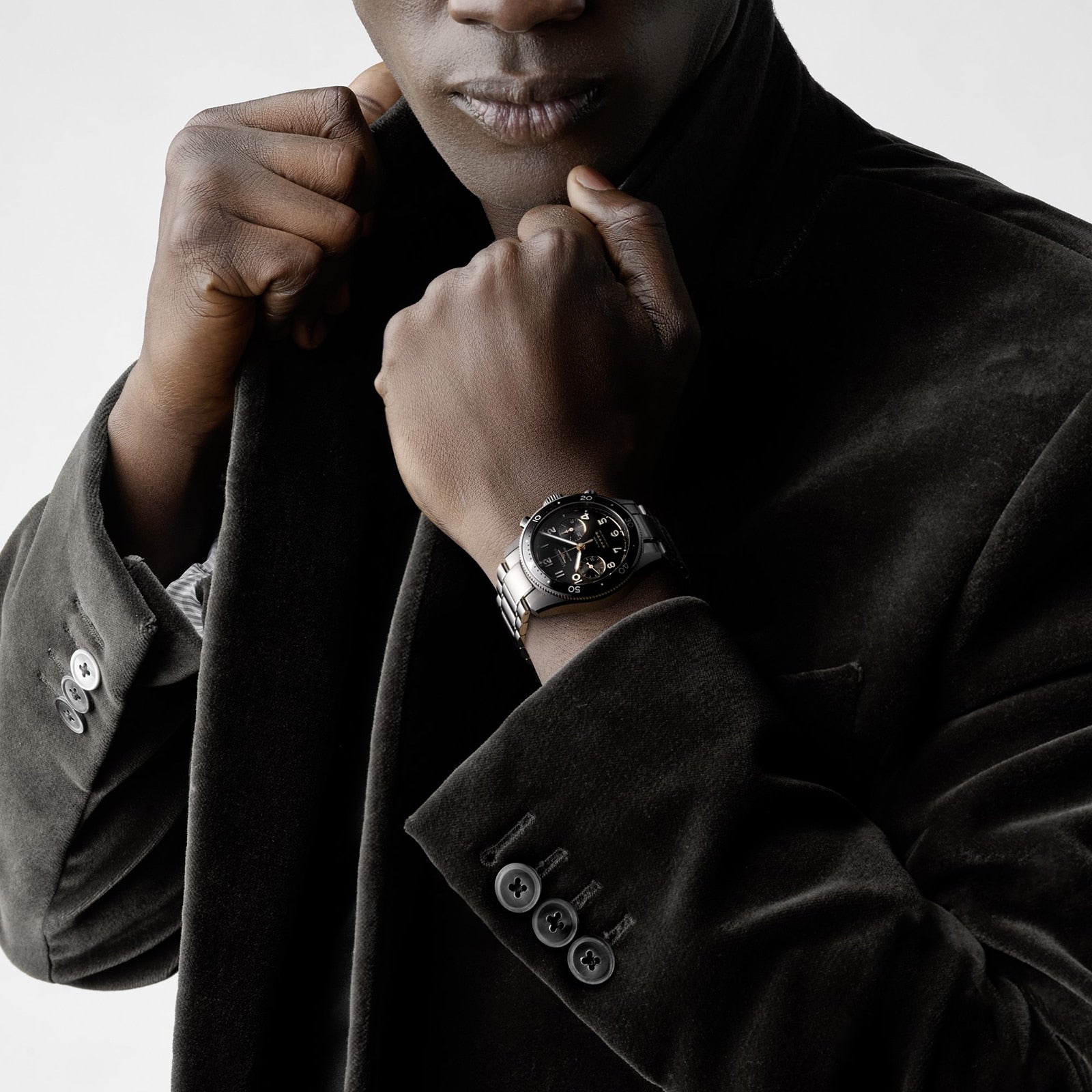 A person in a dark velvet jacket adjusts their collar with one hand, showcasing a Longines Spirit Flyback watch on the other wrist.