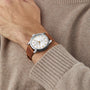 A person wears a beige sweater with a silver Longines Conquest Heritage watch featuring a white dial, gold markers, and a brown leather strap—a Swiss timepiece celebrated for its accuracy.