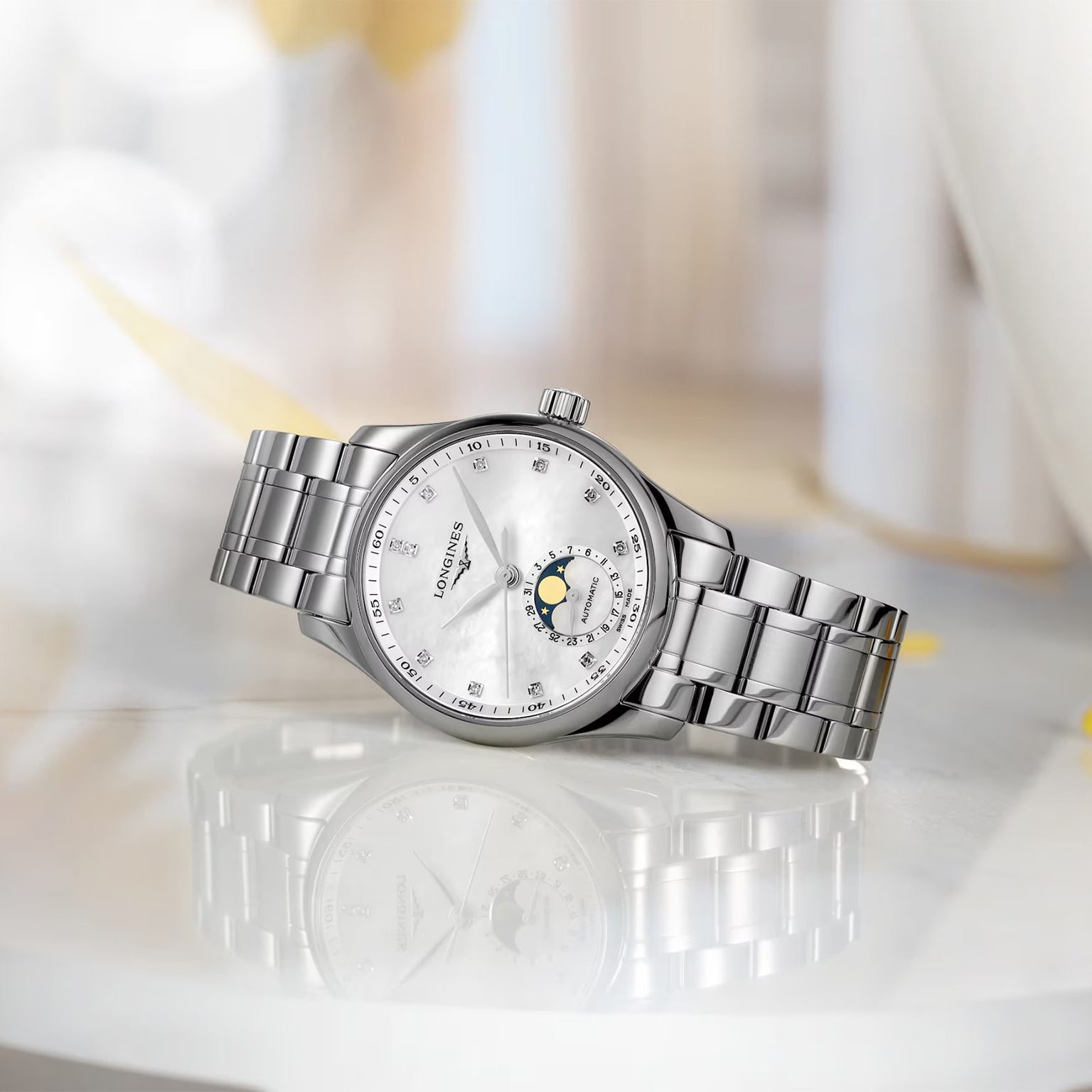 The Longines Master Collection Moonphase, a silver stainless steel watch with a white dial, diamond indexes, date display, and moon phase indicator, is displayed on a reflective surface.
