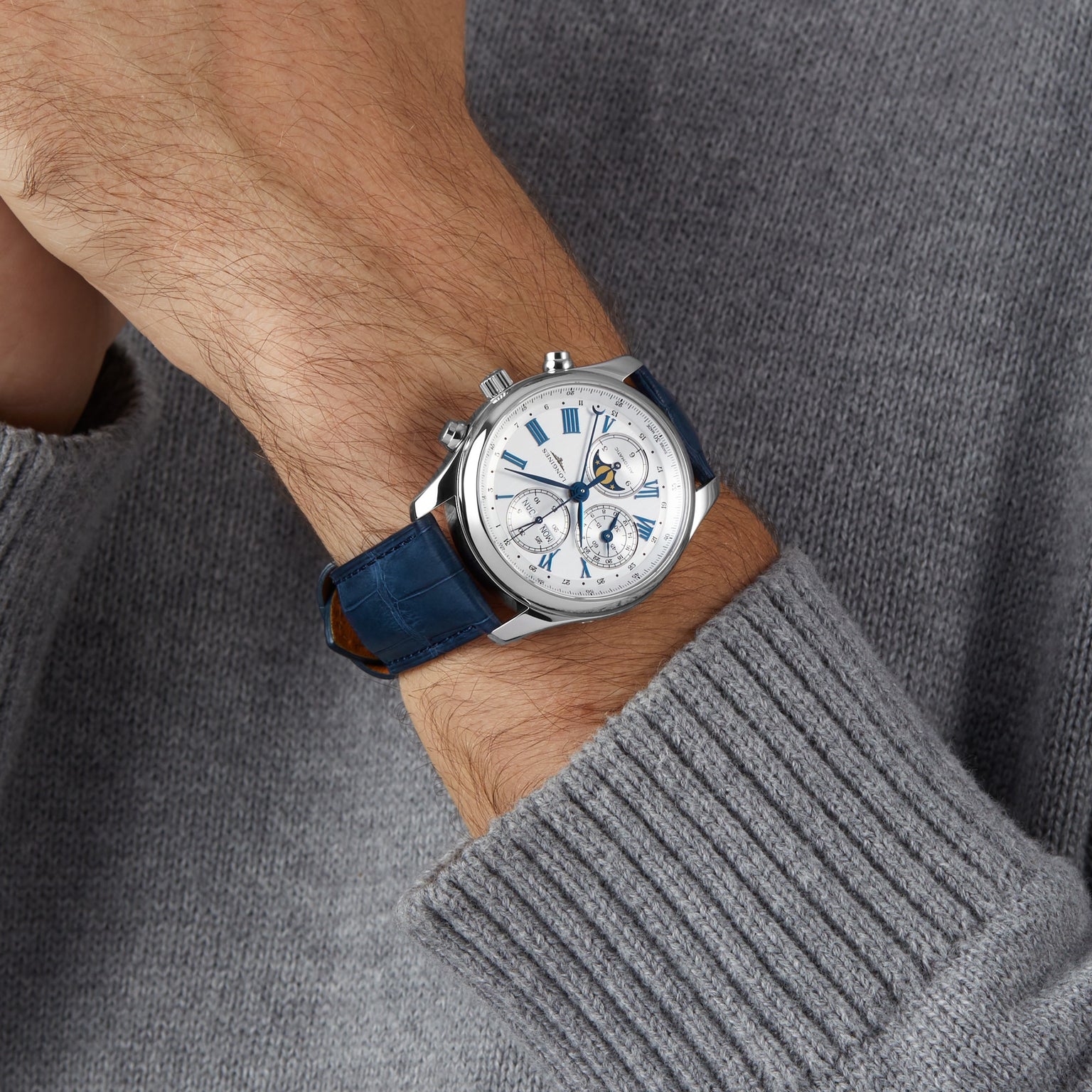 A person wearing a Longines Master Collection Chrono Moonphase silver chronograph with a blue leather strap and a light gray knit sweater.