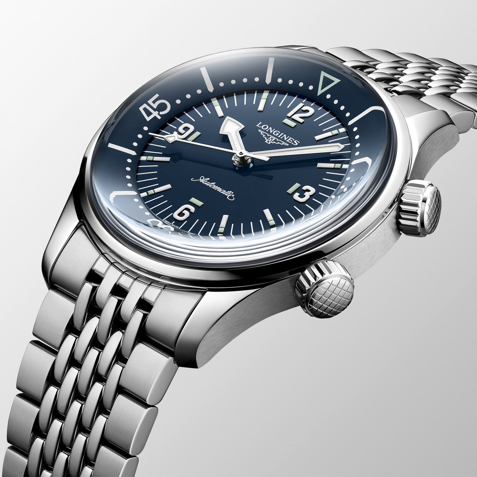 The Longines Legend Diver is a stainless steel automatic watch with a blue dial, luminous markers, two crowns, and a metal link bracelet—shown on a light gray background as a modern interpretation of the classic dive watch.