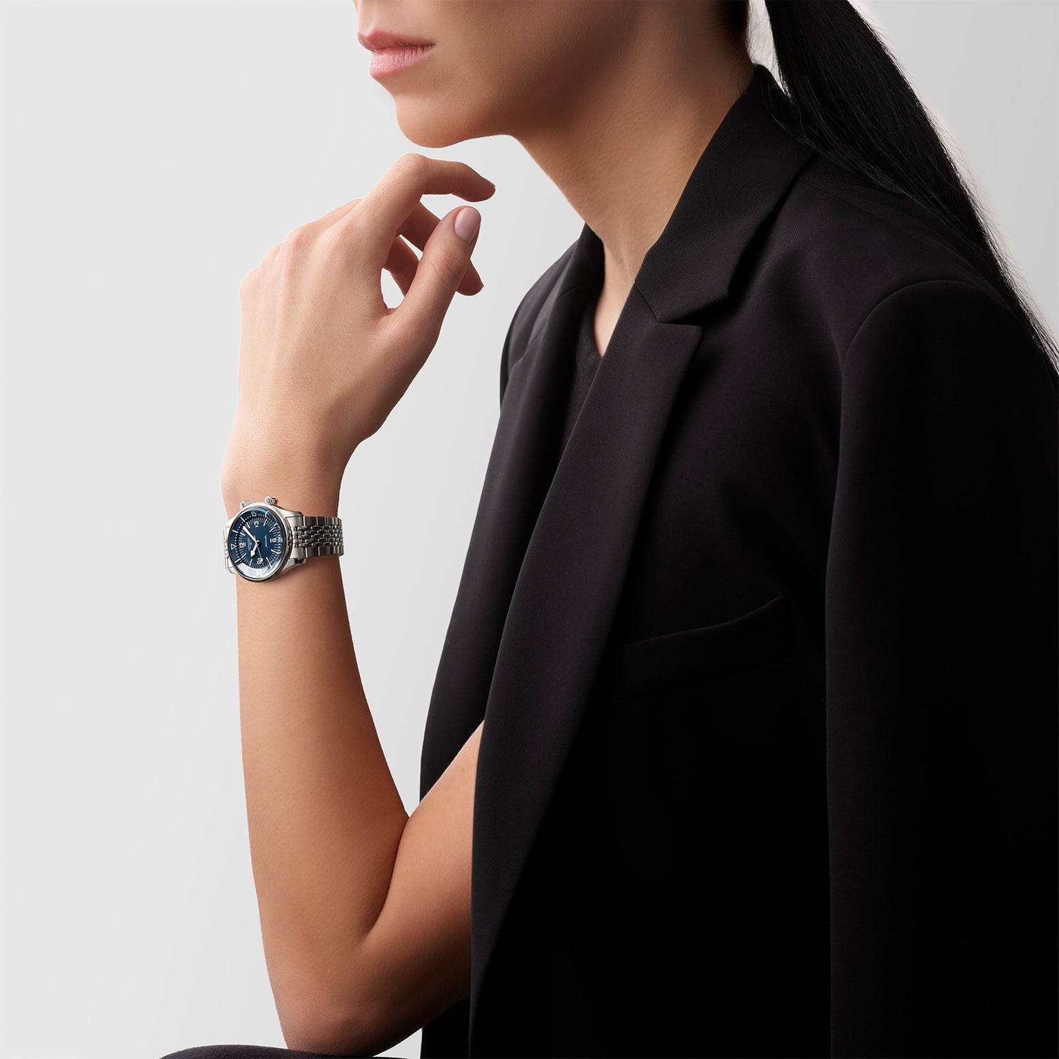 A woman in a black blazer poses in profile, her hand near her chin, wearing the Longines Legend Diver—a vintage-style automatic watch with a dark blue face.