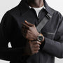 A person in a black zip-up jacket and white shirt adjusts a gray strap across their chest, wearing the Longines Legend Diver watch with a brown leather band.