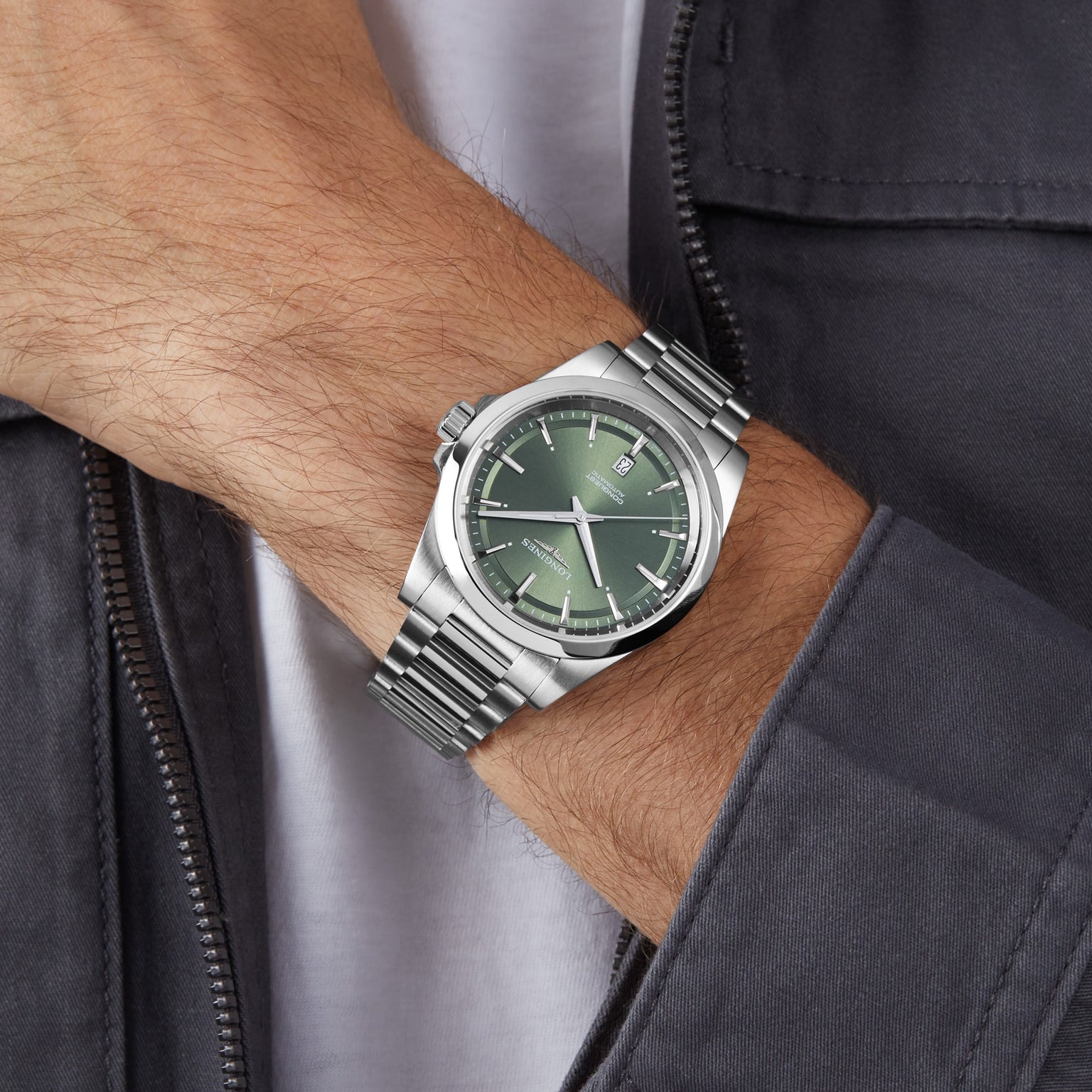 A person wearing a Longines Conquest watch with a green dial, Swiss Super-LumiNova accents, and date display rests their hand near a gray jacket.