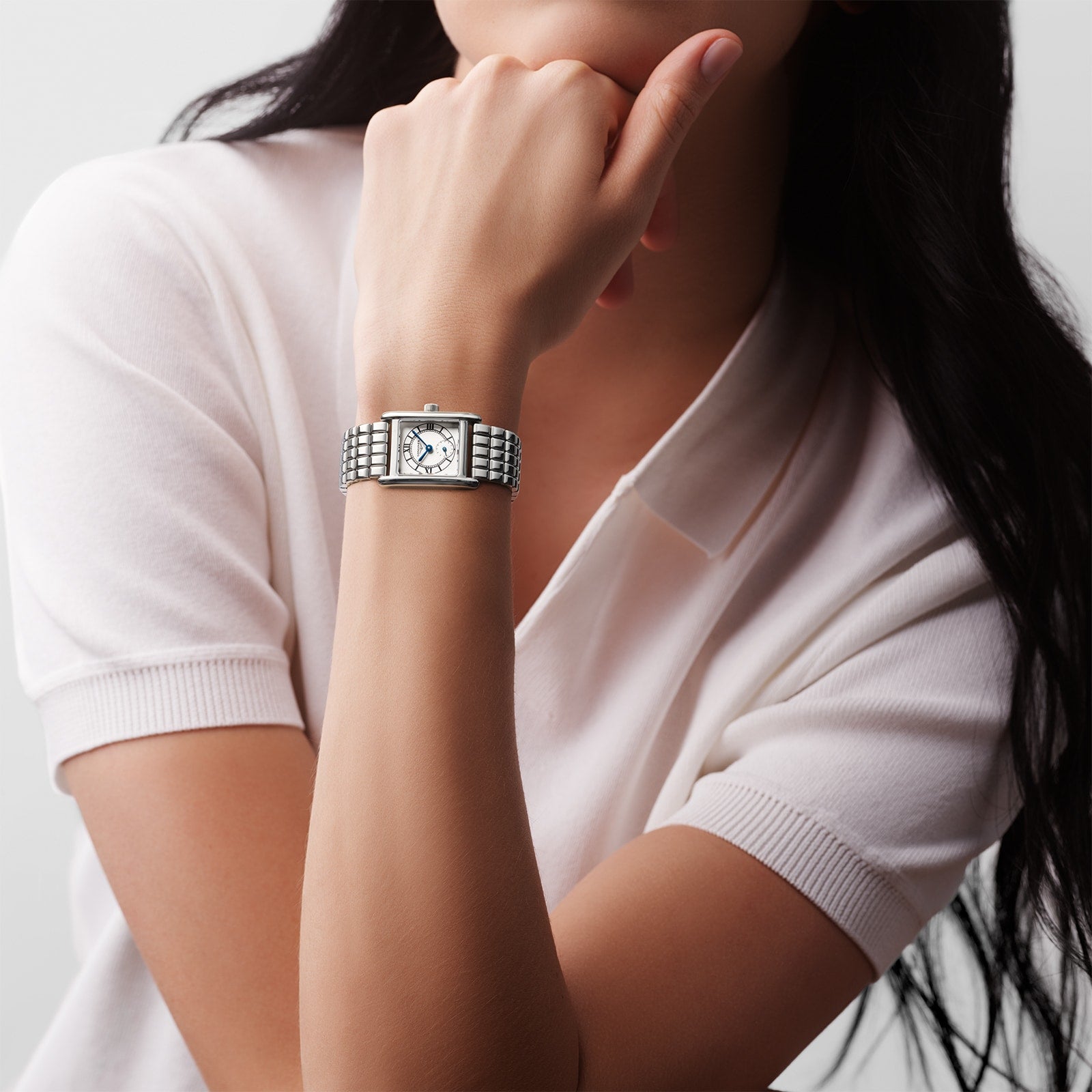 A person wearing a short-sleeved white polo shirt and the Longines Mini DolceVita watch has their hand near their chin, perfectly showcasing contemporary sophistication.