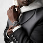 A person in a black suit and white hoodie showcases the silver Longines Conquest Heritage Central Power Reserve wristwatch with Swiss SuperLumiNova® coating on their left wrist against a neutral background.