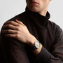 A person in a black turtleneck and translucent long-sleeve shirt displays a Longines Conquest Heritage Central Power Reserve wristwatch with a stainless steel case and dark strap on their left wrist.