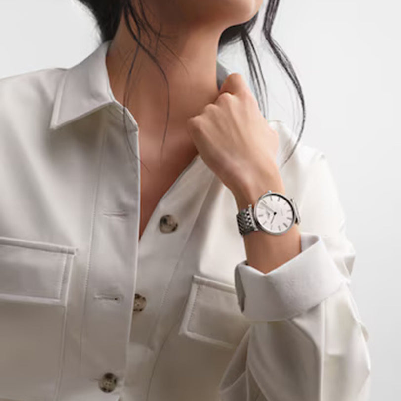 A person, exuding classic elegance, rests their chin on their hand while wearing a silver La Grande Classique de LONGINES watch by Longines and a light-colored button-up shirt against a plain background.