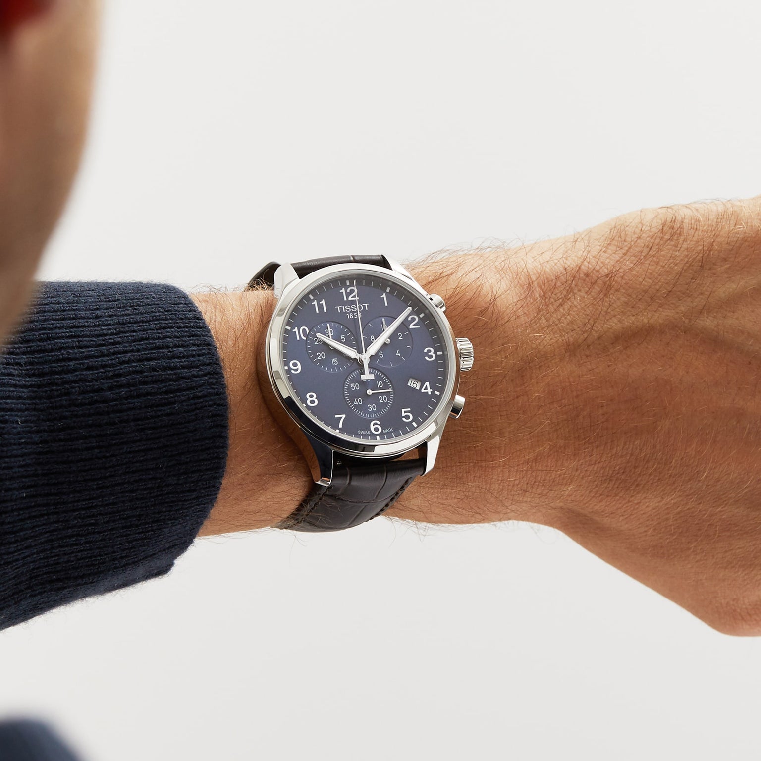 A wrist wearing a Tissot Chrono XL Classic with a dark leather strap, navy blue dial, and silver casing, set against a plain light background.