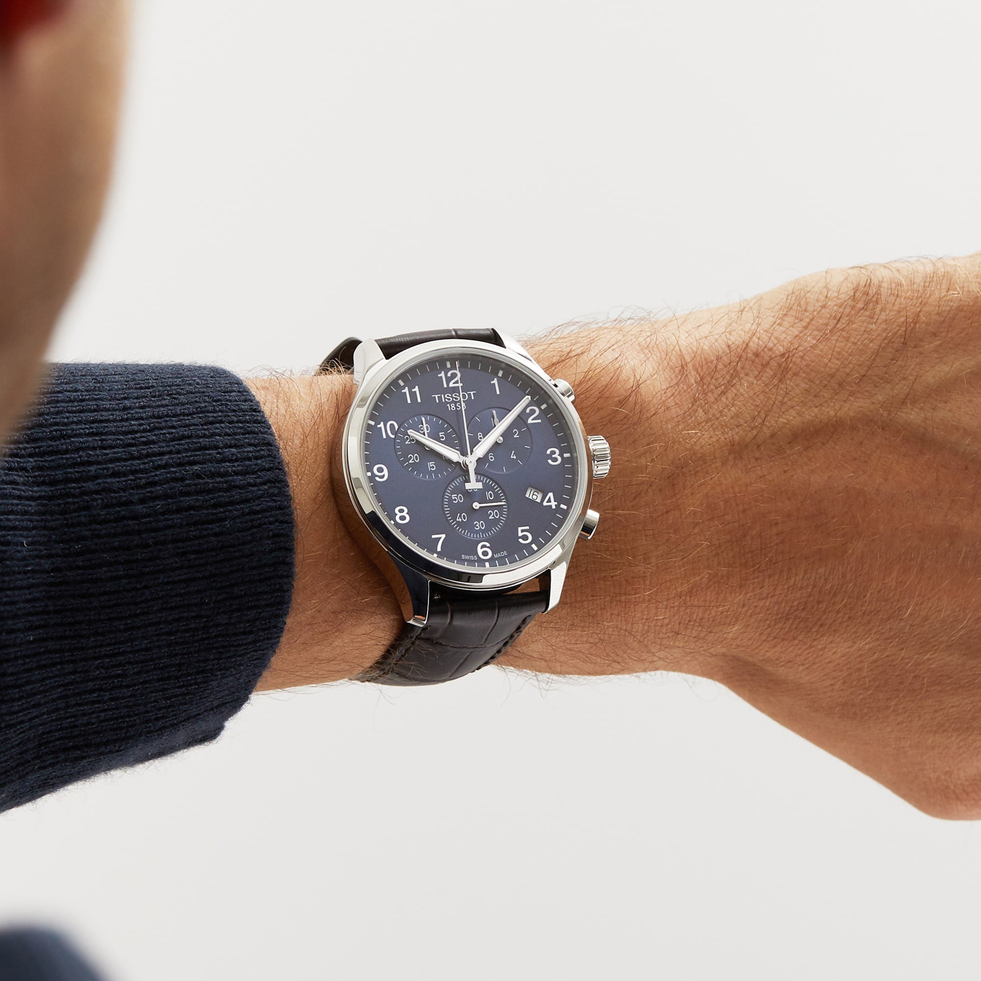 A wrist wearing a Tissot Chrono XL Classic with a dark leather strap, navy blue dial, and silver casing, set against a plain light background.