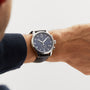 A wrist wearing a Tissot Chrono XL Classic with a dark leather strap, navy blue dial, and silver casing, set against a plain light background.