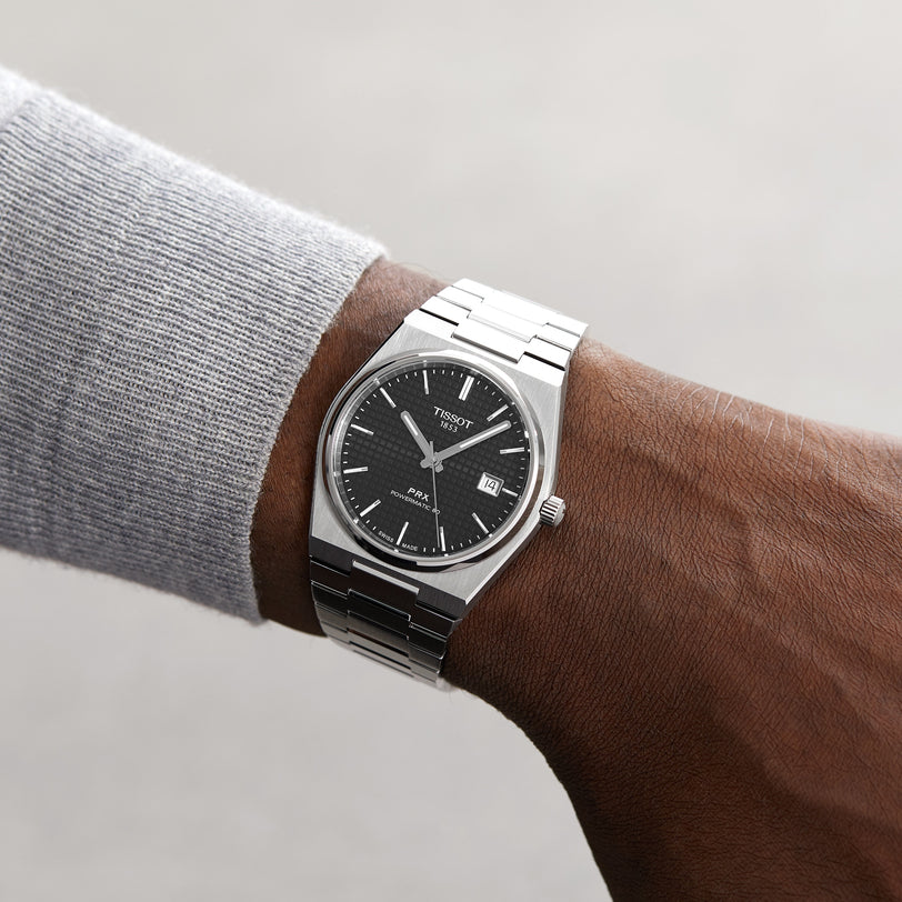 A person wearing the Tissot PRX Powermatic 80 with a black dial, stainless steel case and metal band checks the time—about 1:49. The elegant design also features a date window.
