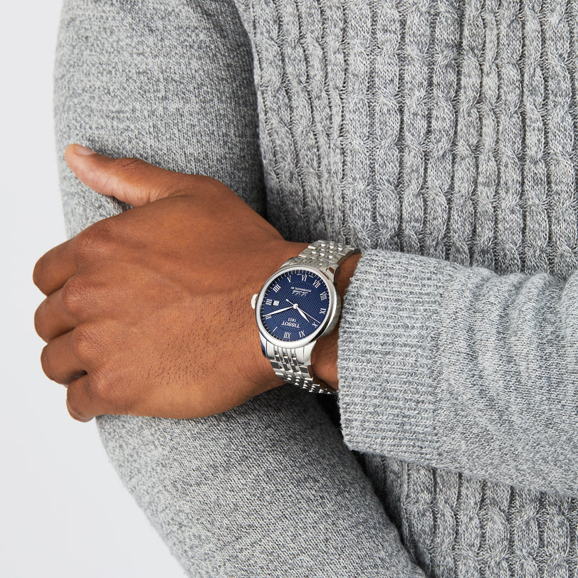 A person in a textured gray sweater displays a wrist with the Tissot Le Locle Powermatic 80, a silver watch featuring a blue dial, Roman numerals, and renowned Swiss precision.