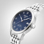 The Tissot Le Locle Powermatic 80, a silver wristwatch with a blue textured dial, Roman numeral hour markers, and metal link bracelet, is displayed against a plain gradient background.
