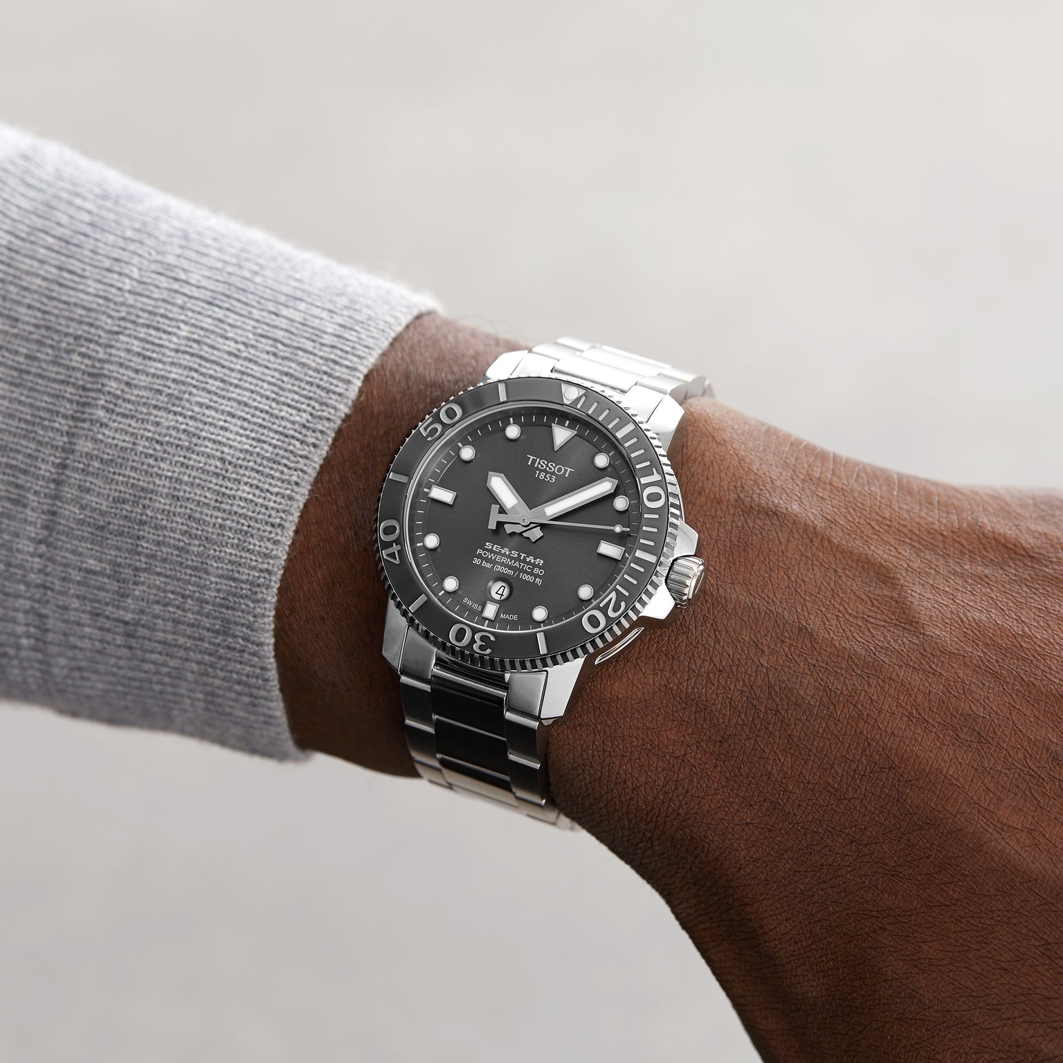 A person wearing a Tissot Seastar 1000 Powermatic 80 wristwatch with a black face and metal band on their left wrist, shown against a plain background.