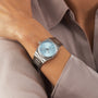 A person wearing a Tissot PRX silver wristwatch with a light blue face, dressed in a light gray long-sleeve shirt.