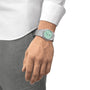 A person in a white shirt and grey pants wears a Tissot PRX watch with a mint green face, highlighting its sleek design on their left wrist.