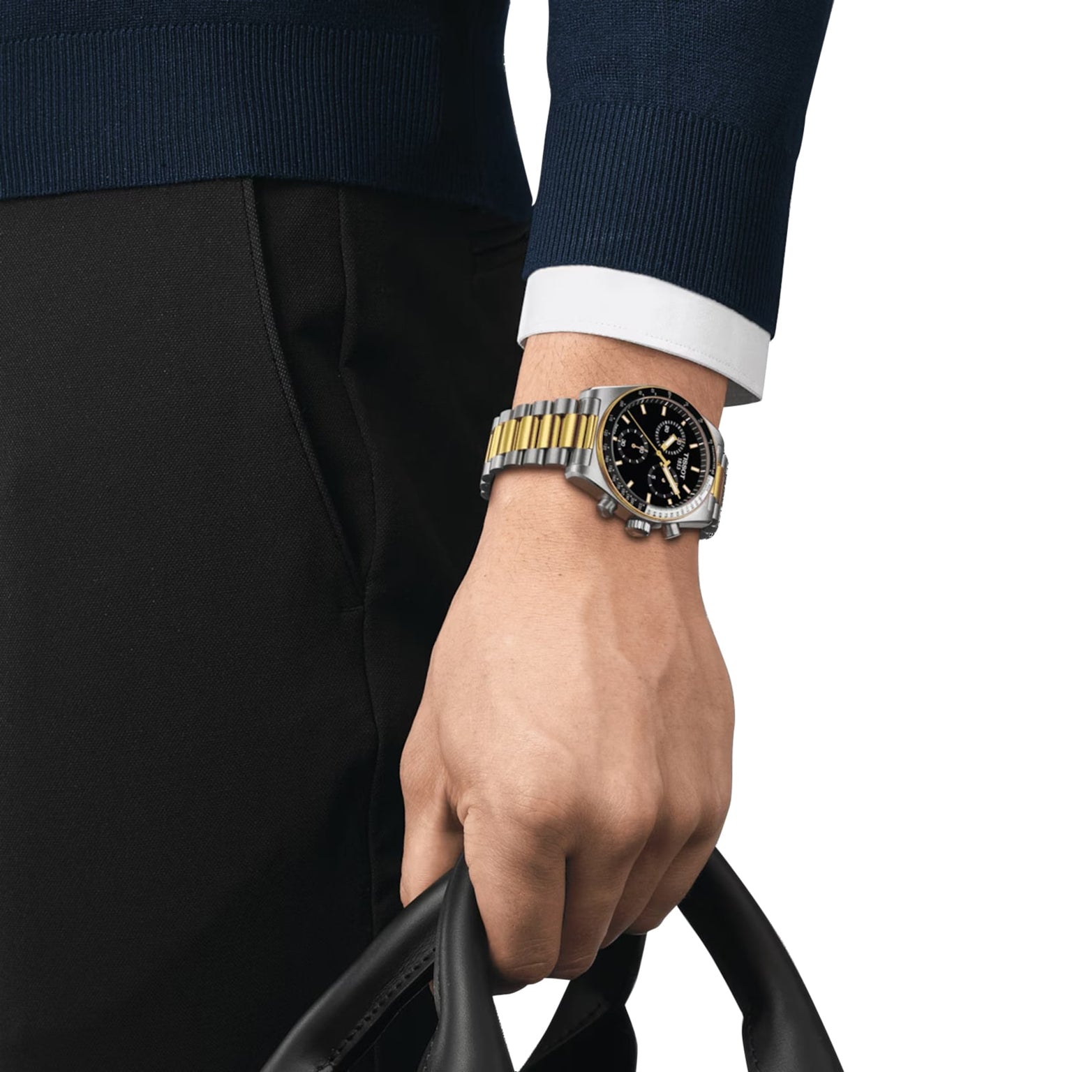 A person in a dark sweater and pants holds a black bag; on their left wrist is a Tissot PR516 Chronograph with a silver and gold finish, black dial, and quartz movement.