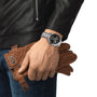 A person in a black leather jacket and jeans holds brown leather gloves and wears a silver Tissot PR516 Chronograph watch.