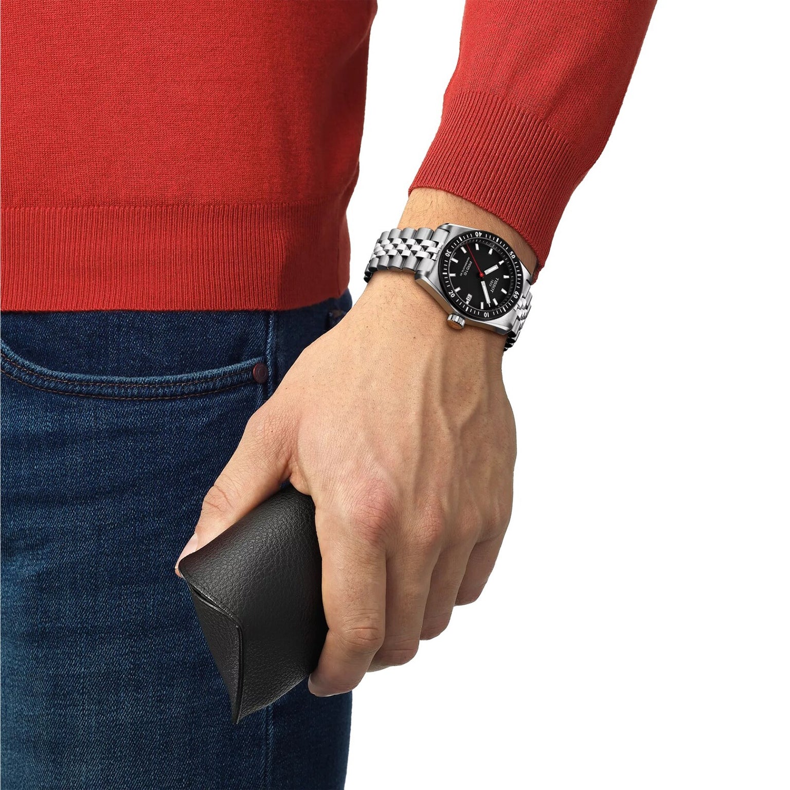 A person in a red sweater and jeans wears a Tissot PR516 Powermatic 80 silver wristwatch and holds a black case.
